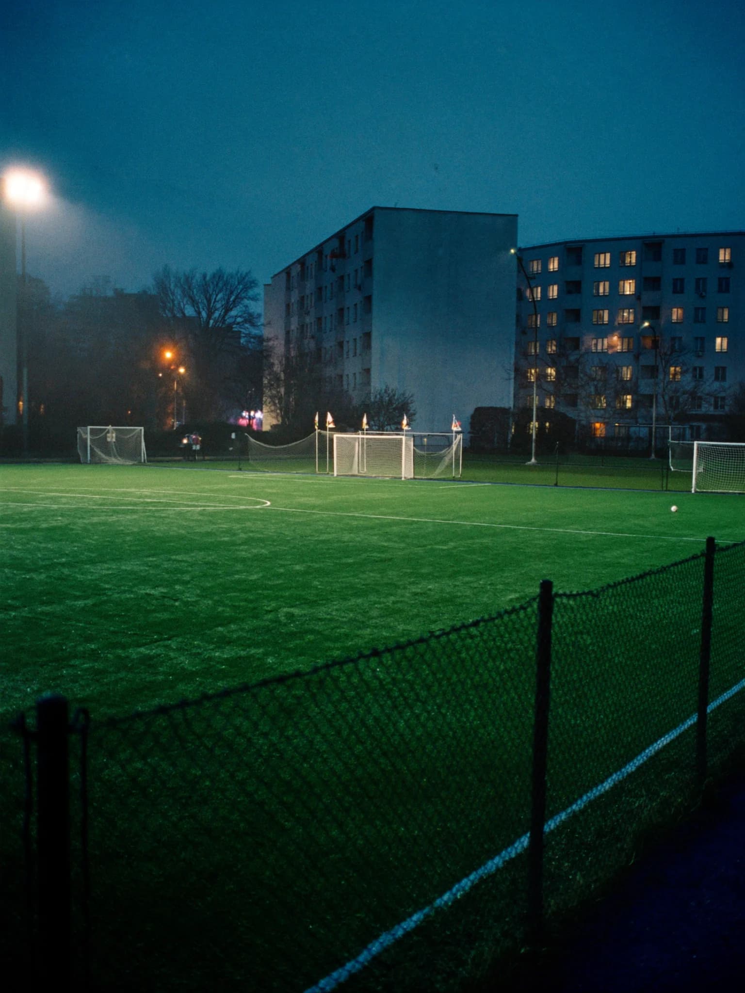 A floodlit five-a-side pitch at night between apartment blocks, seen through a chain-link fence